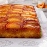 A rectangular Upside-Down Buttermilk Peach Cake with caramelized peach slices on top sits on a white patterned platter. Two glass bottles and a small bowl of peach slices are blurred in the background.