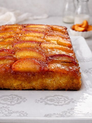 A rectangular Upside-Down Buttermilk Peach Cake with caramelized peach slices on top sits on a white patterned platter. Two glass bottles and a small bowl of peach slices are blurred in the background.