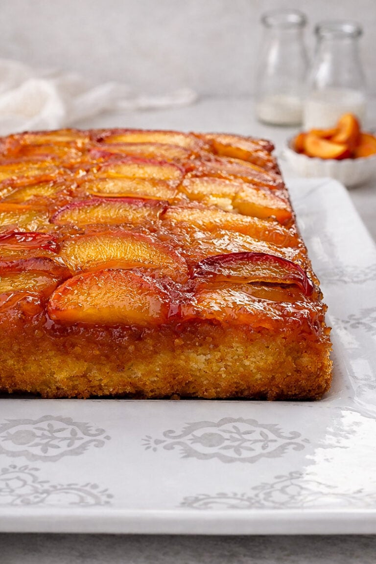 A rectangular Upside-Down Buttermilk Peach Cake with caramelized peach slices on top sits on a white patterned platter. Two glass bottles and a small bowl of peach slices are blurred in the background.