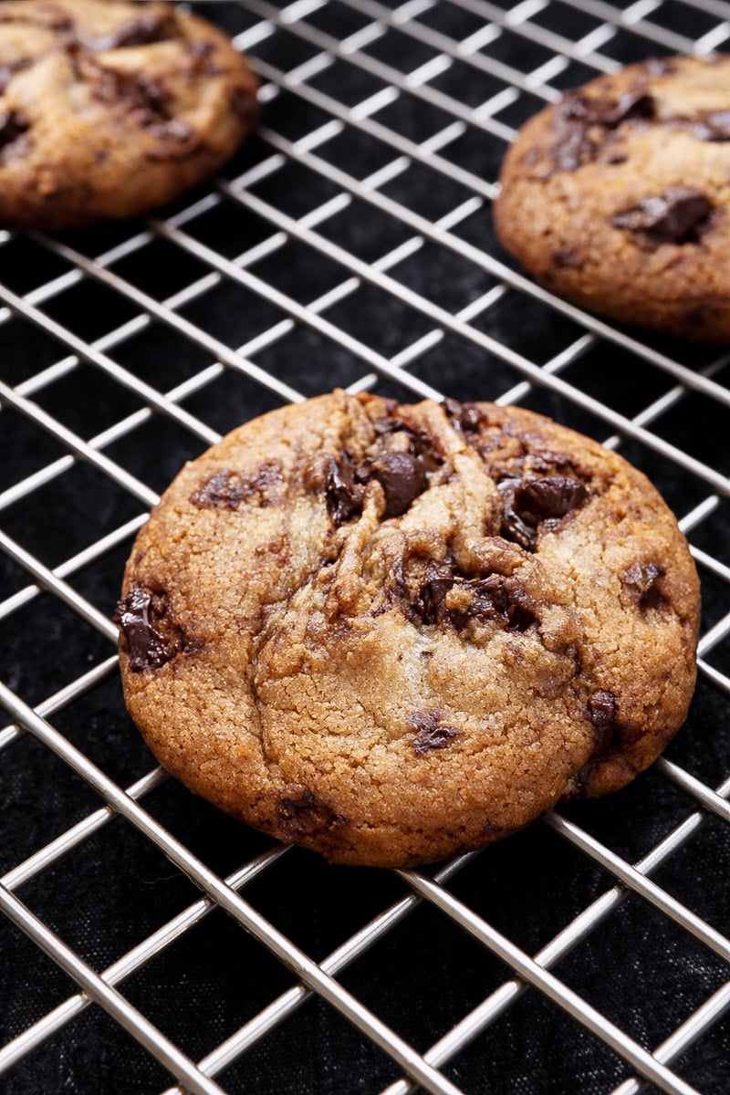 A close-up of The Best Browned Butter and Molasses Chocolate Chip Cookies cooling on a metal wire rack, with more cookies visible in the background. The cookies appear golden brown with melted chocolate chunks.