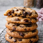 A stack of The Best Browned Butter and Molasses Chocolate Chip Cookies sits on a cooling rack, the top cookie partially eaten. Two glass jars, one with brown sugar and one with chocolate chips, blur in the background.