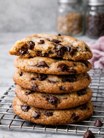 A stack of The Best Browned Butter and Molasses Chocolate Chip Cookies sits on a cooling rack, the top cookie partially eaten. Two glass jars, one with brown sugar and one with chocolate chips, blur in the background.
