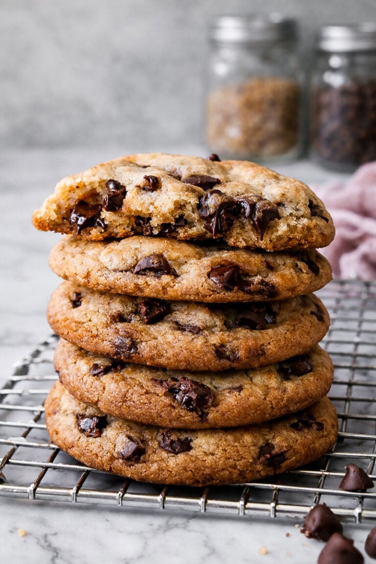 A stack of The Best Browned Butter and Molasses Chocolate Chip Cookies sits on a cooling rack, the top cookie partially eaten. Two glass jars, one with brown sugar and one with chocolate chips, blur in the background.
