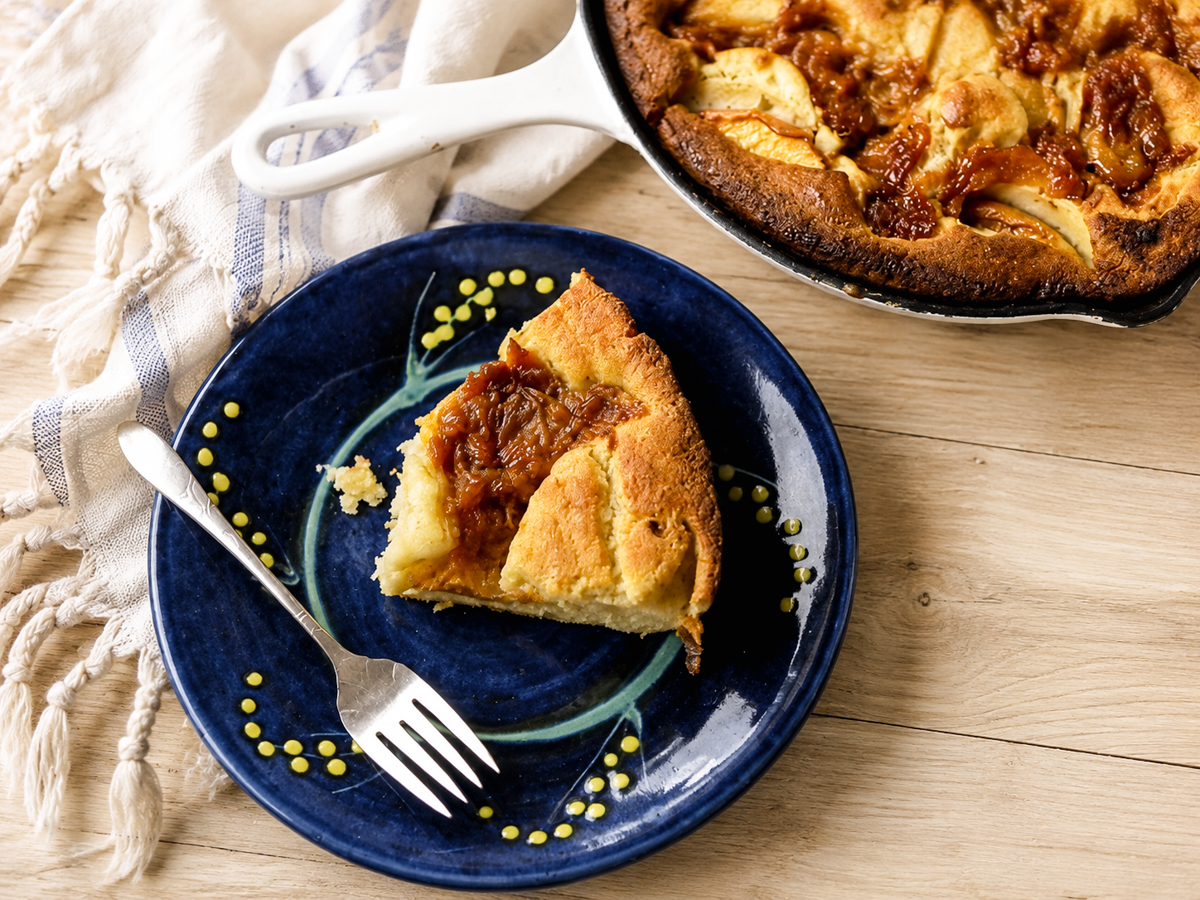 A slice of golden baked apple Dutch baby pancake sits on a blue plate with a fork, next to a skillet—perfect for making Skillet Peach Cobbler—containing the rest of the pancake on a wooden table with a cream and blue striped towel.