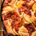 A closeup of Skillet Peach Cobbler baked in a cast iron skillet, showing golden-brown crust, caramelized edges, and juicy peach slices on top. The skillet is resting on a wooden surface.