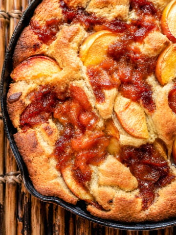 A closeup of Skillet Peach Cobbler baked in a cast iron skillet, showing golden-brown crust, caramelized edges, and juicy peach slices on top. The skillet is resting on a wooden surface.