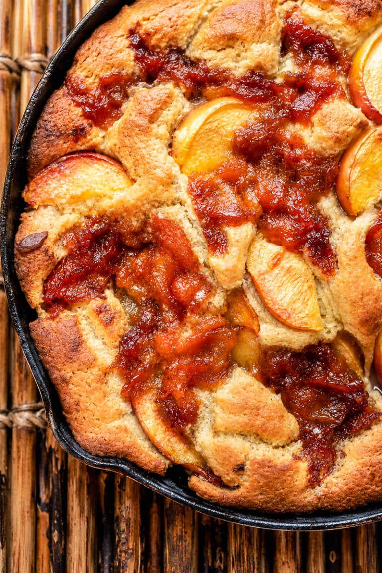 A closeup of Skillet Peach Cobbler baked in a cast iron skillet, showing golden-brown crust, caramelized edges, and juicy peach slices on top. The skillet is resting on a wooden surface.
