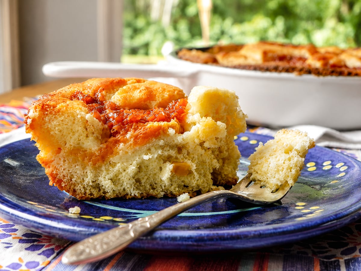 A close-up of a slice of golden brown Skillet Peach Cobbler on a blue plate, with a piece cut and lifted by a fork. The background shows a baking dish and a bright, sunlit window.