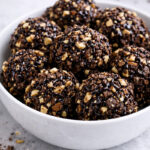 A white bowl filled with round chocolate and nut clusters drizzled with chocolate sauce, set on a light gray surface. A few berries are blurred in the background.