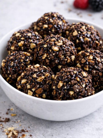 A white bowl filled with round chocolate and nut clusters drizzled with chocolate sauce, set on a light gray surface. A few berries are blurred in the background.