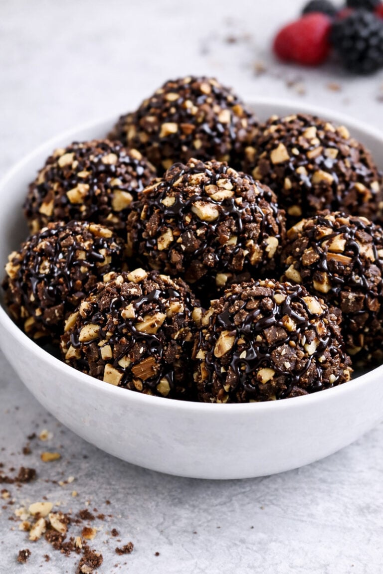 A white bowl filled with round chocolate and nut clusters drizzled with chocolate sauce, set on a light gray surface. A few berries are blurred in the background.