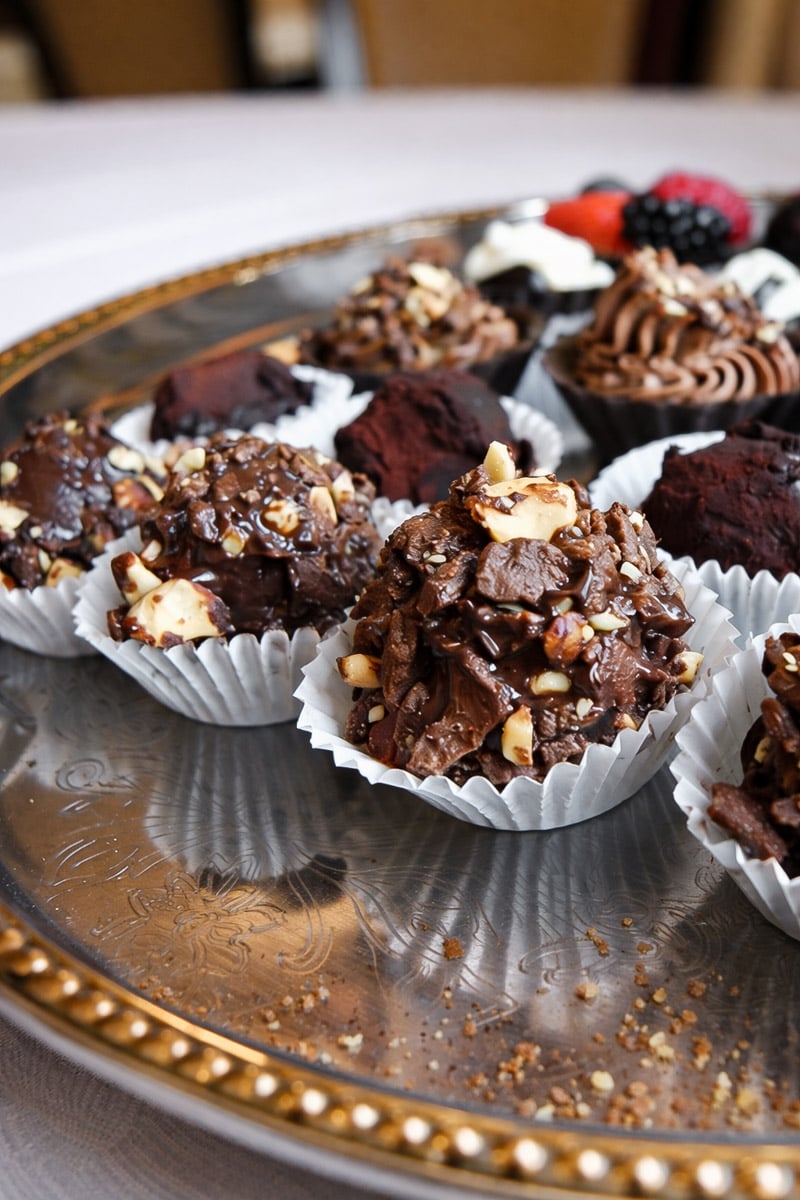 A close-up of assorted chocolate desserts and pastries on a silver tray, featuring chocolate clusters with nuts in white paper cups and other decorated treats in the background.