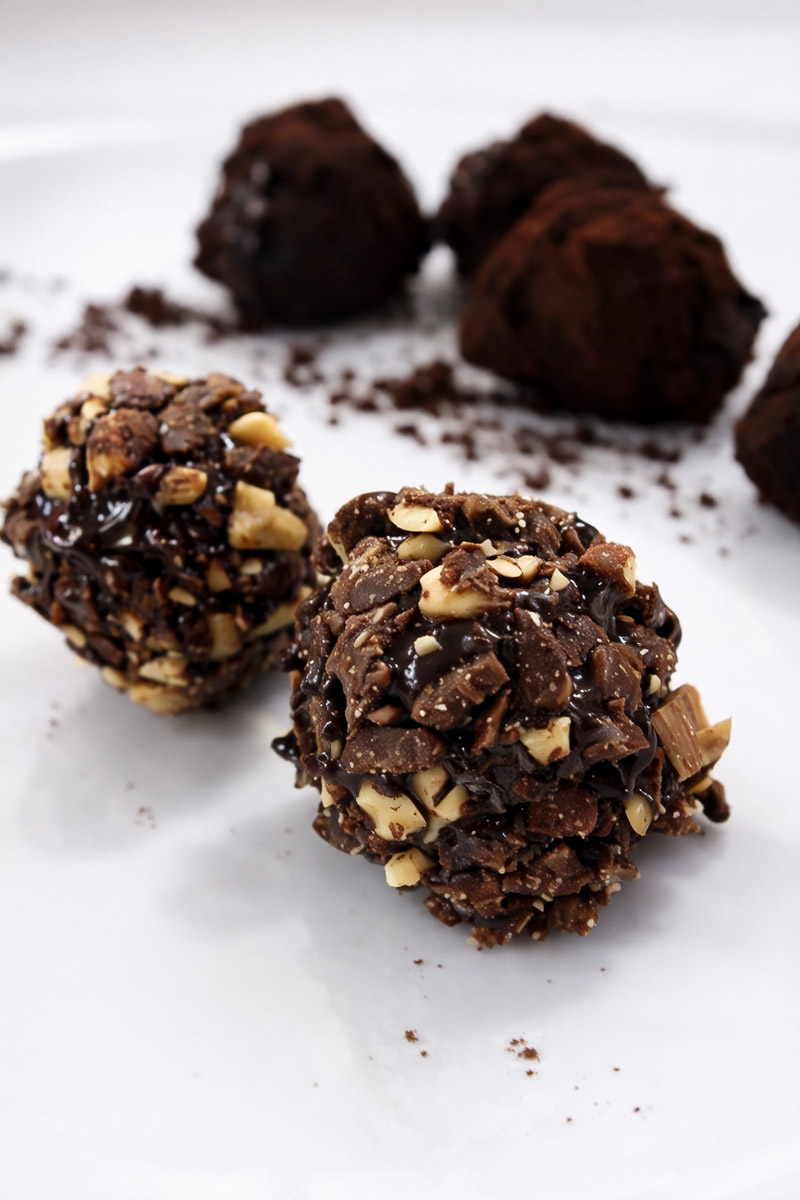 Close-up of chocolate truffles on a white plate; two truffles are coated with chopped nuts and drizzled with chocolate, while others in the background are dusted with cocoa powder.
