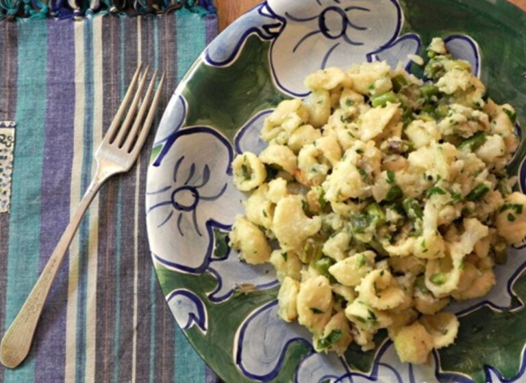 A plate of shell pasta mixed with chopped green vegetables sits on a colorful floral plate next to a striped placemat and a silver fork.