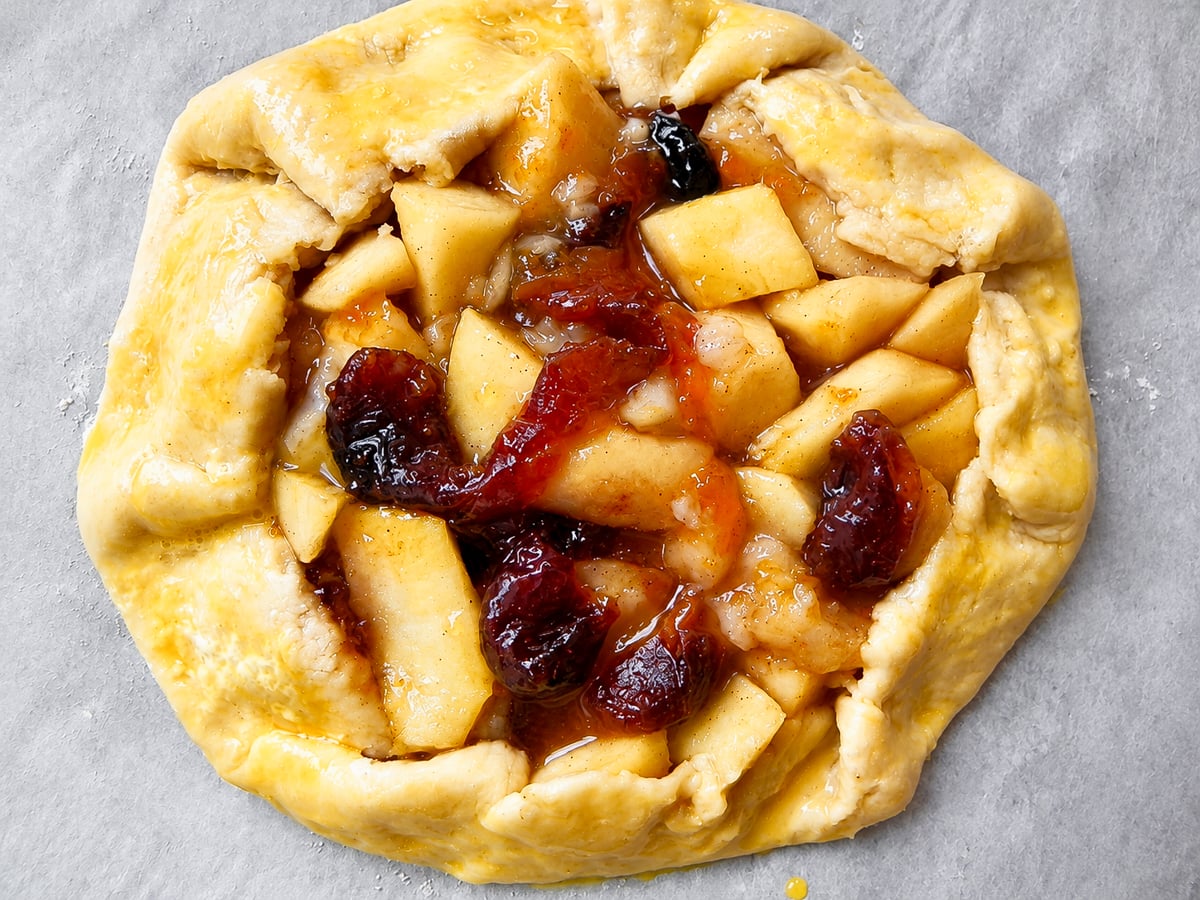 Unbaked apple and dried fruit Crostata Autunnale with a golden, egg-washed crust on parchment paper.