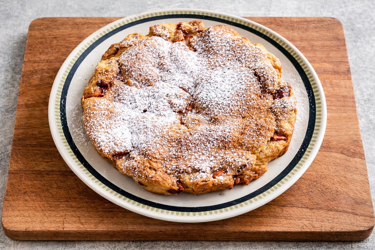 A rustic pear cake dusted with powdered sugar sits on a white plate with a black rim, placed on a wooden cutting board against a light gray background.