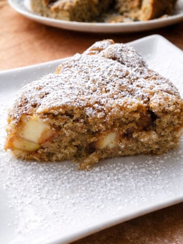 A slice of apple cake with visible apple pieces, dusted with powdered sugar, sits on a white plate. In the background, a rustic pear cake rests on another plate, slightly blurred.