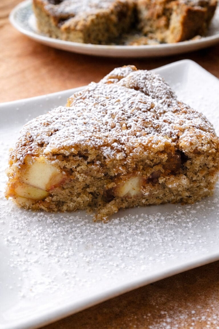 A slice of apple cake with visible apple pieces, dusted with powdered sugar, sits on a white plate. In the background, a rustic pear cake rests on another plate, slightly blurred.