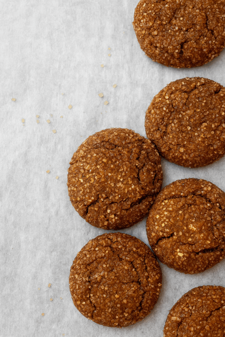 Five round, golden-brown Ginger Sugar Cookies with a sugar-coated, cracked surface are arranged on white parchment paper, with a few sparkling sugar granules scattered nearby.