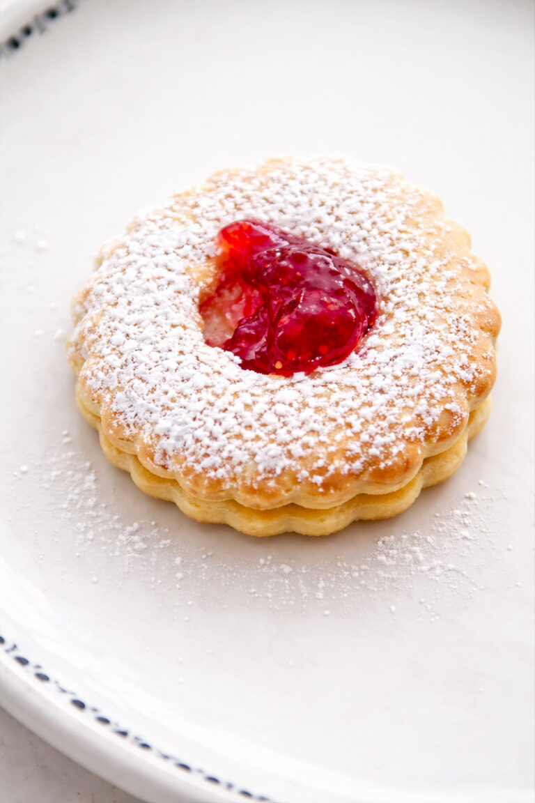 A round, scalloped cookie dusted with powdered sugar, featuring a center filled with bright red jam, is placed on a white plate.