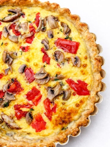 A close-up of a baked Torta Salata with a golden, flaky crust, topped with sliced mushrooms, red bell peppers, and melted cheese on a white background.