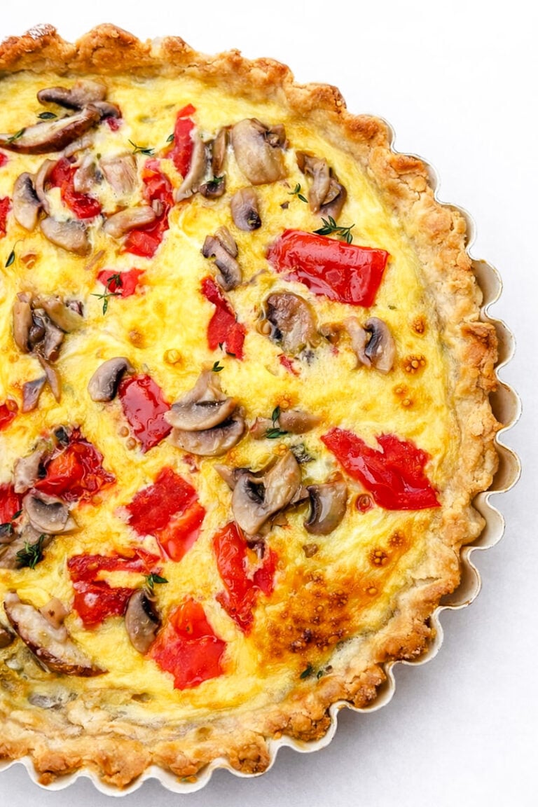 A close-up of a baked Torta Salata with a golden, flaky crust, topped with sliced mushrooms, red bell peppers, and melted cheese on a white background.