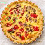 A freshly baked torta salata in a fluted pie dish, topped with sliced mushrooms, red bell peppers, and herbs, sits on parchment paper. The golden crust and creamy filling make this mixed vegetable galette irresistible.