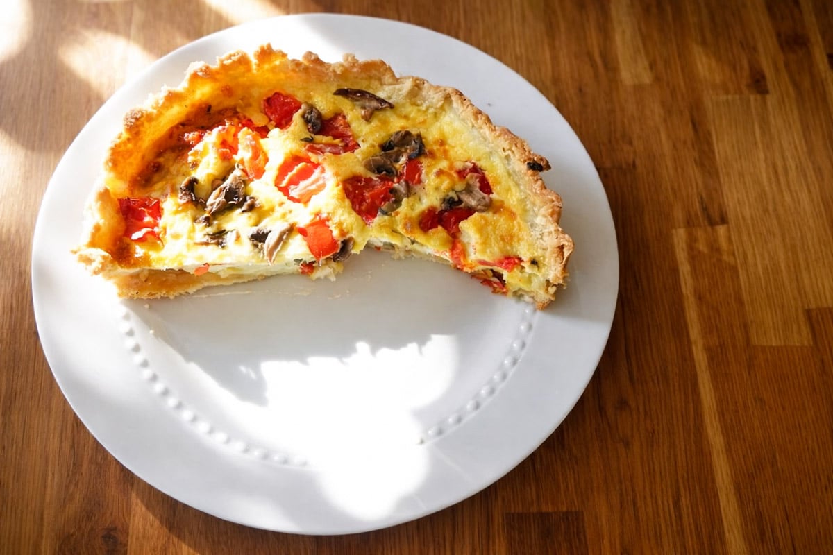 A partially eaten Torta Salata with a golden crust sits on a white plate atop a wooden table, sunlight casting gentle shadows across the mixed vegetable galette and the inviting scene.