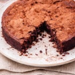 A round chocolate cake dusted with cocoa powder sits on a plate. A large wedge has been cut out and removed, showing the moist, dense interior. The plate rests on a piece of burlap atop a light surface.