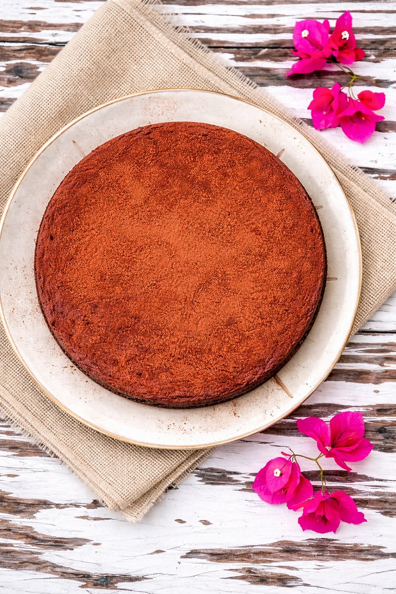 chocolate-and-chestnut-cake-torta-al-cioccolato-e-le-castagne-2 A round chocolate cake dusted with cocoa powder sits on a white plate atop a beige cloth. Pink flowers are scattered nearby on a rustic white wooden table.