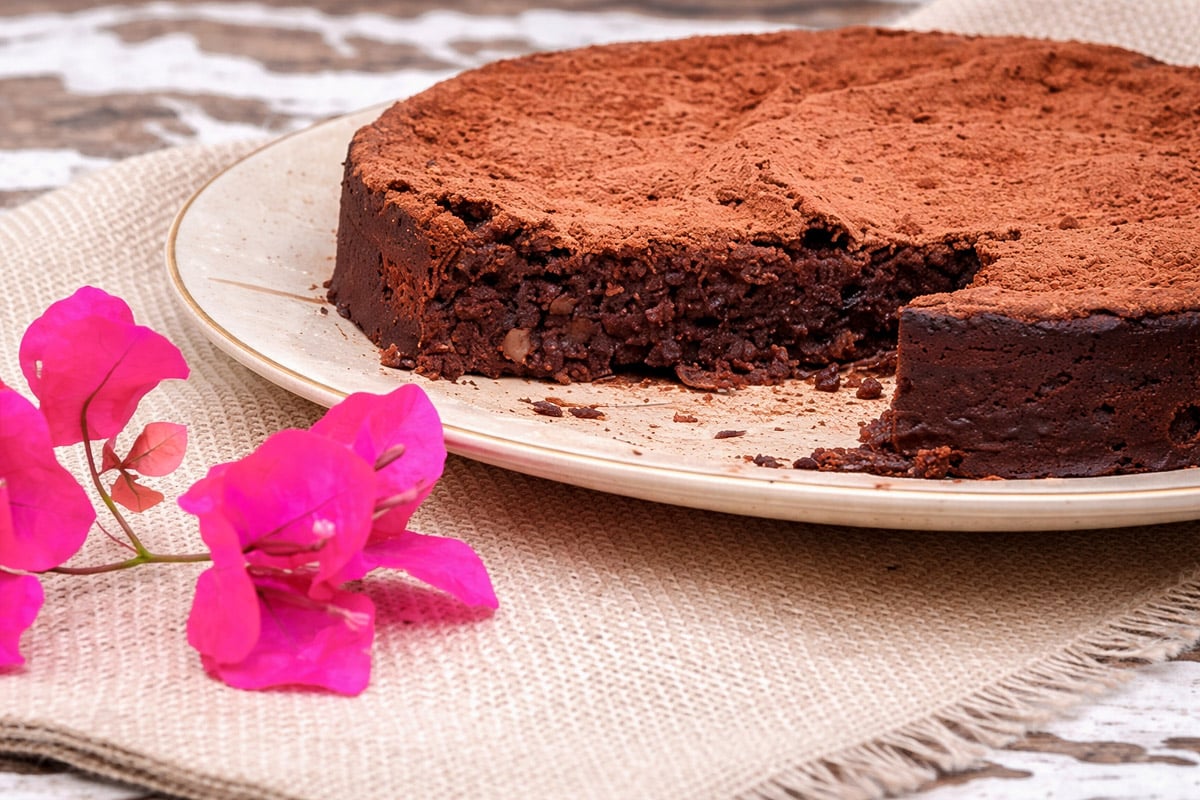 chocolate-and-chestnut-cake-torta-al-cioccolato-e-le-castagne-6 A round, rich chocolate cake with a slice missing sits on a plate beside bright pink flowers and a woven cloth on a rustic wooden surface. The cake is dusted with cocoa powder.