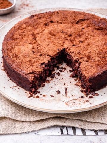 A round chocolate cake dusted with cocoa powder sits on a plate. A large wedge has been cut out and removed, showing the moist, dense interior. The plate rests on a piece of burlap atop a light surface.