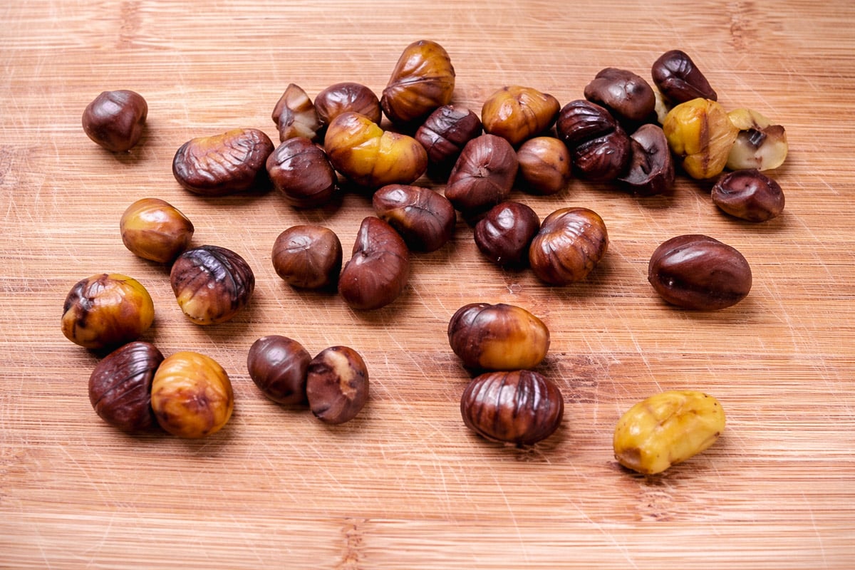 A group of roasted chestnuts, some with shells partially peeled, scattered on a wooden cutting board.