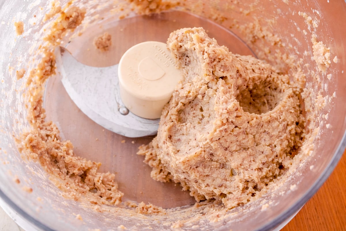 A food processor bowl containing a thick mixture being blended, with some of the dough-like paste gathered near the blade.