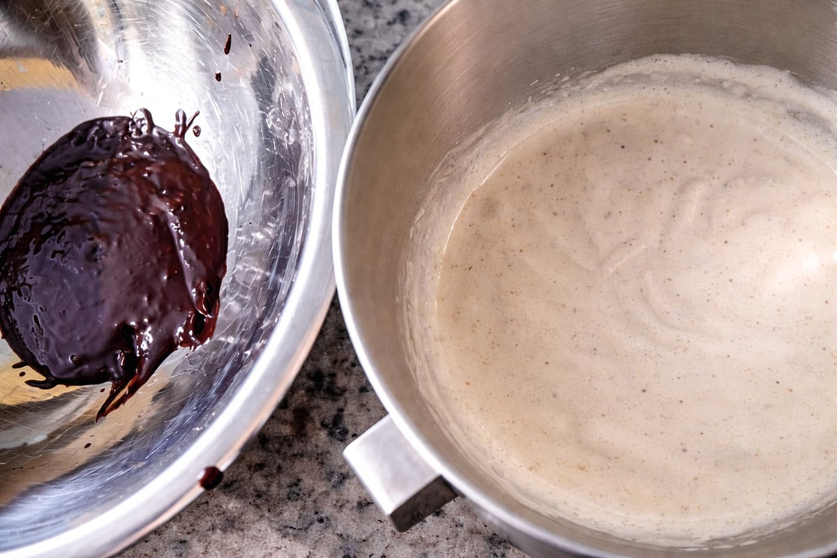 Two metal mixing bowls on a countertop: one contains a small amount of melted chocolate, the other holds a light, frothy batter, possibly for baking or dessert preparation.
