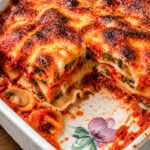 A baked Eggplant Lasagne-Lasagne di Melanzane in a white ceramic dish with a floral design. A large portion is cut out, revealing layers of pasta, spinach, cheese, tomato sauce, mushrooms, and golden crispy top.