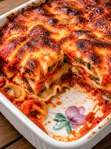 A baked Eggplant Lasagne-Lasagne di Melanzane in a white ceramic dish with a floral design. A large portion is cut out, revealing layers of pasta, spinach, cheese, tomato sauce, mushrooms, and golden crispy top.