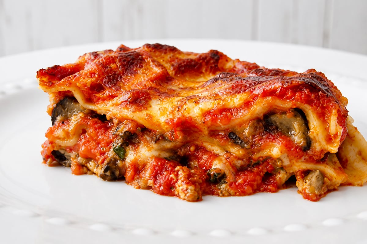 A close-up of a slice of Eggplant Lasagne-Lasagne di Melanzane with layers of pasta, tomato sauce, melted cheese, and vegetables on a white plate.
