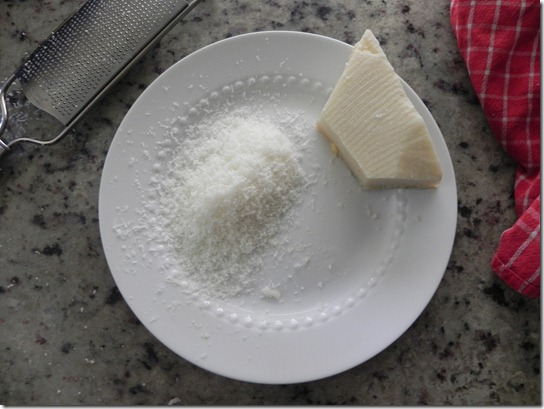 A wedge of hard cheese and a pile of freshly grated cheese sit on a white plate next to a metal grater, with a red kitchen towel on a speckled countertop.