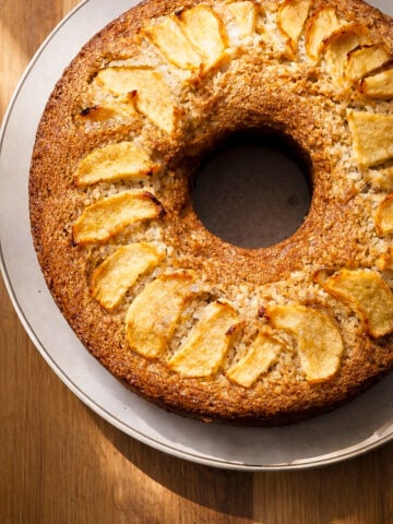 A close-up of a golden-brown Vegan Apple Ring Cake-Ciambella di Mele on a white plate, topped with neatly arranged slices of baked apple. The cake sits on a wooden table, with light casting gentle shadows.