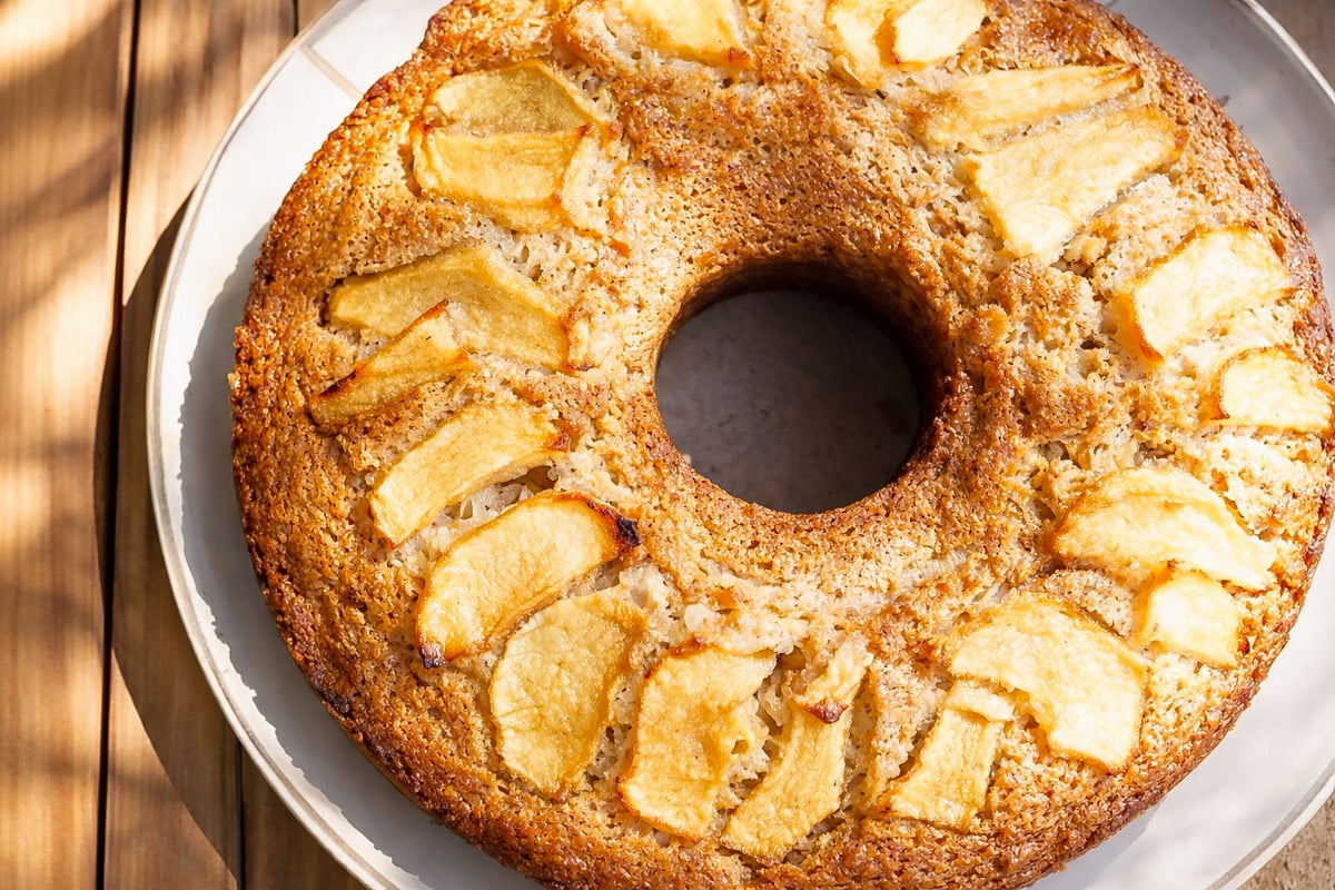 A round Vegan Apple Ring Cake-Ciambella di Mele with a hole in the center, topped with baked apple slices, sits on a white plate on a wooden surface. The cake has a golden-brown crust and a moist texture.