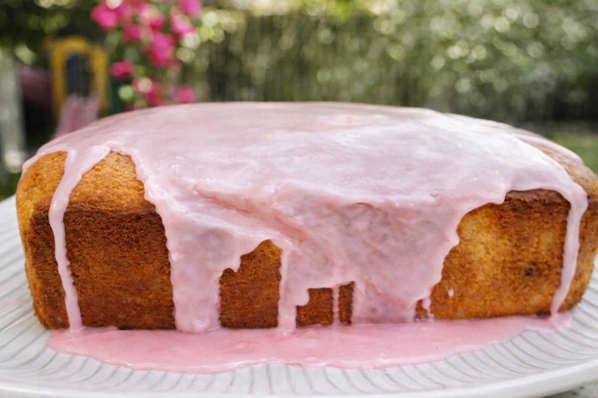 A rectangular Meyer Lemon Bread with Blood Orange Glaze features golden brown sides and pink icing that drips down the edges, set on a white plate with a blurred garden background.