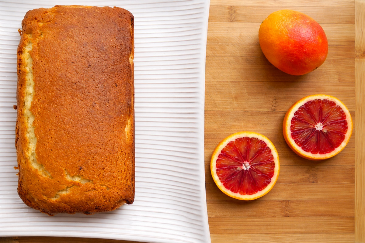 A golden-brown Meyer Lemon Bread with Blood Orange Glaze sits on a white textured plate next to a whole blood orange and two blood orange halves on a wooden surface.