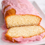 A loaf of Meyer Lemon Bread with Blood Orange Glaze, topped with glossy pink icing, sits on a white plate. The inside of the cake appears moist and fluffy, making it an irresistible treat.