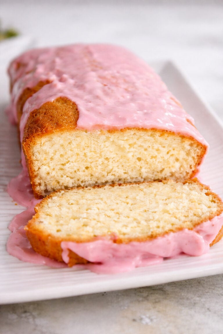 A loaf of Meyer Lemon Bread with Blood Orange Glaze, topped with glossy pink icing, sits on a white plate. The inside of the cake appears moist and fluffy, making it an irresistible treat.