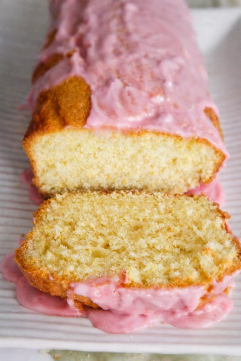A loaf of Meyer Lemon Bread with Blood Orange Glaze, partially sliced to reveal its soft, moist interior. One slice is cut and lies in front of the loaf on a white rectangular plate.