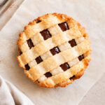 A round pie with a golden lattice crust and visible chocolate filling sits on parchment paper, with a beige cloth napkin nearby.