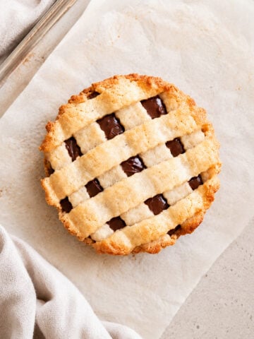 A round pie with a golden lattice crust and visible chocolate filling sits on parchment paper, with a beige cloth napkin nearby.