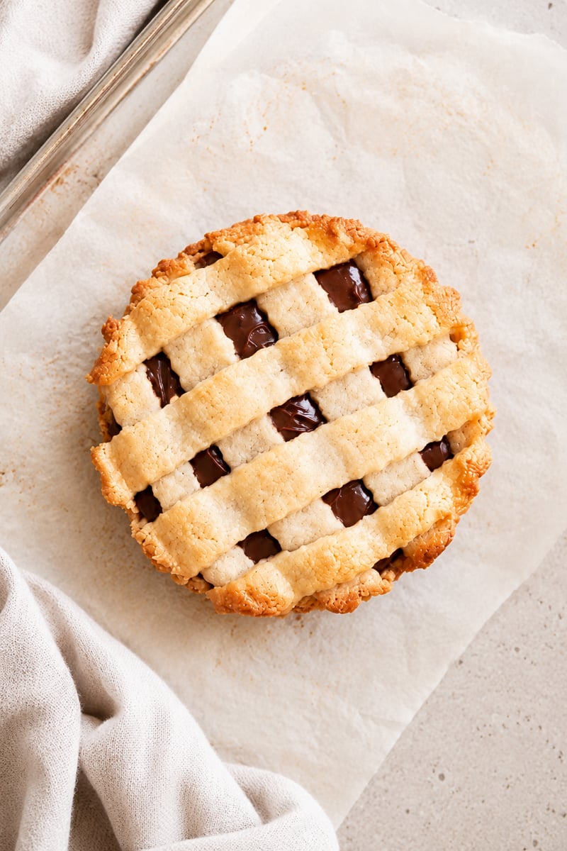A round pie with a golden lattice crust and visible chocolate filling sits on parchment paper, with a beige cloth napkin nearby.