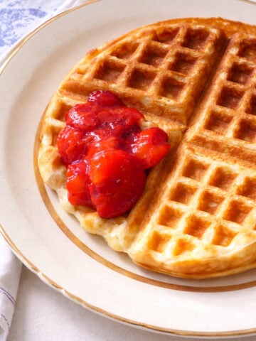 A golden brown yeast waffle on a beige-rimmed plate, topped with a spoonful of bright red strawberry compote, sits on a white tablecloth.